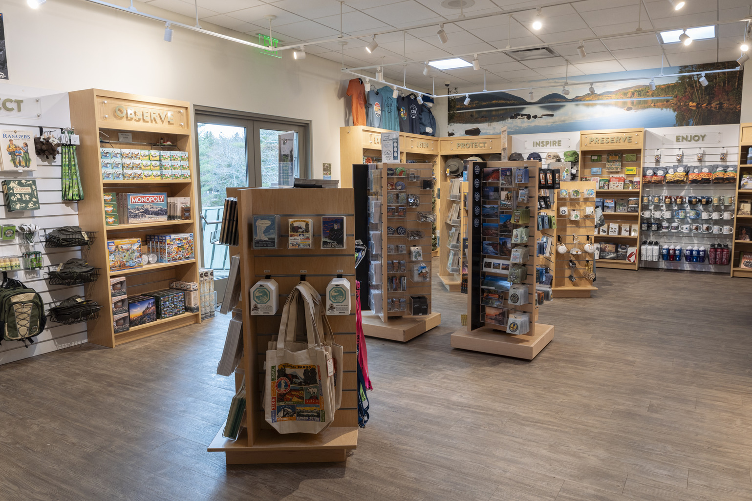 Interior of Acadia's Park Store with wooden shelves and racks displaying games, clothing, and souvenirs, under signs encouraging