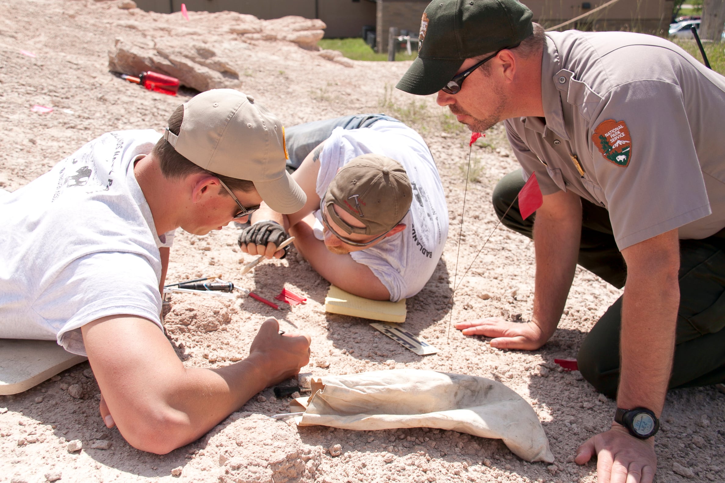 Two paleontologists examining a dig site using brushes, pens, and other tools of the trade. One uniformed park ranger observes n