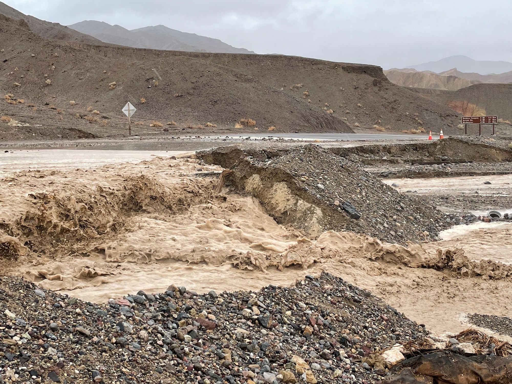 Muddy floodwater flowing through a desert landscape across a paved road causing undercutting on the downhill side.