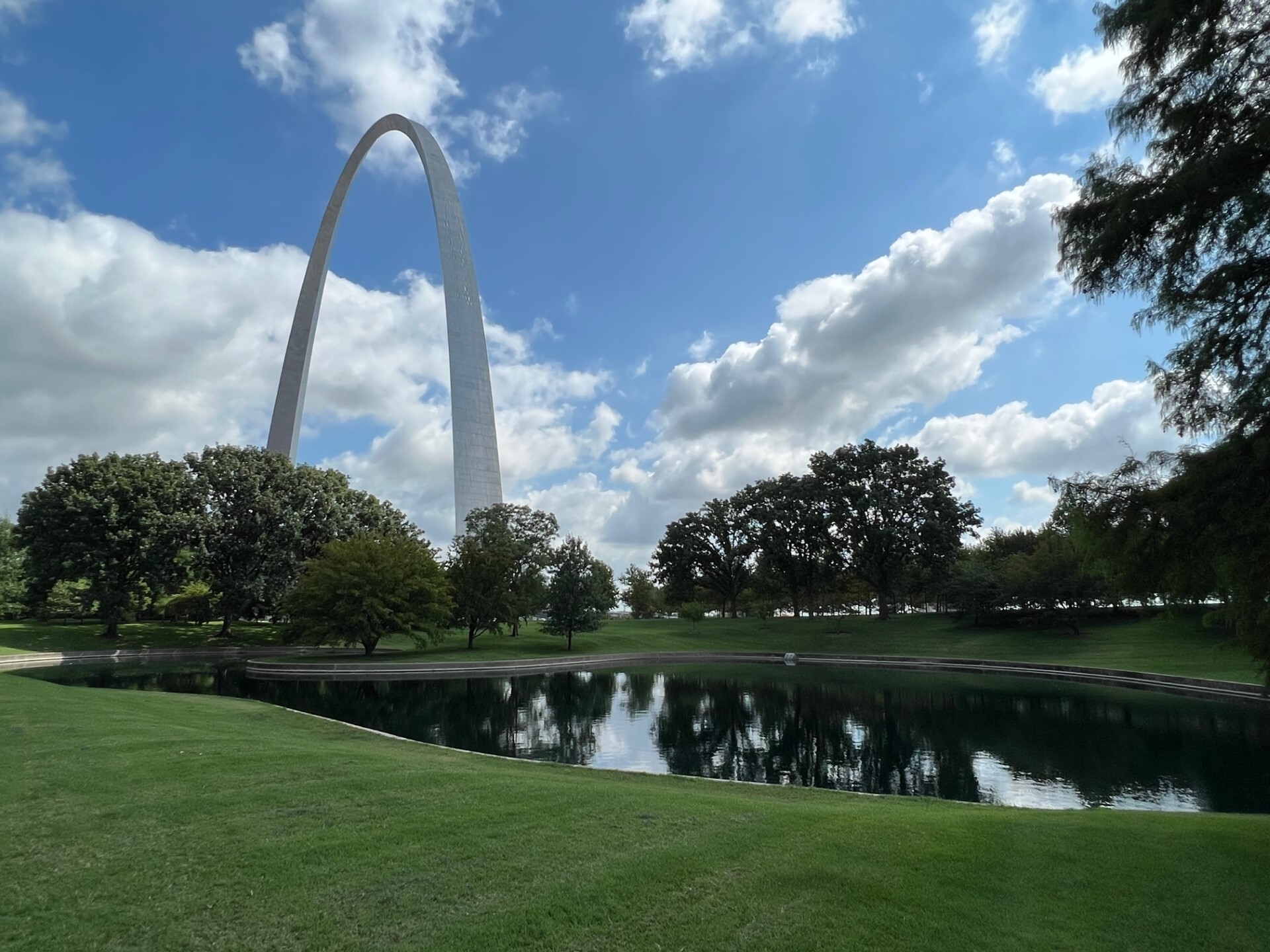 Green grass and the south pond are in the foreground, a line of green trees runs through the center of the image. The Gateway Ar