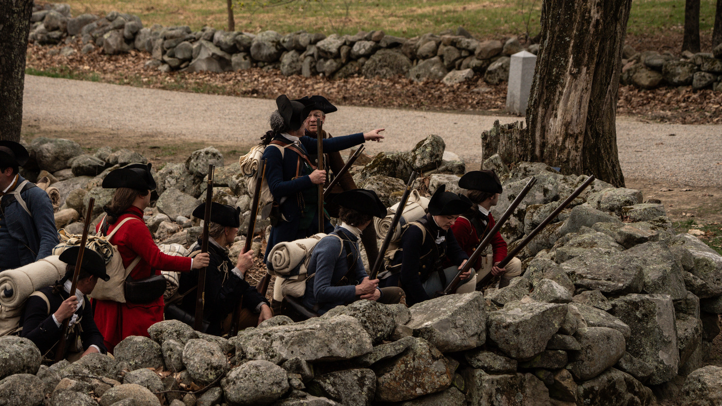 A group of men dressed as Provincial Minute Men hide behind a stone wall that borders a dirt road.