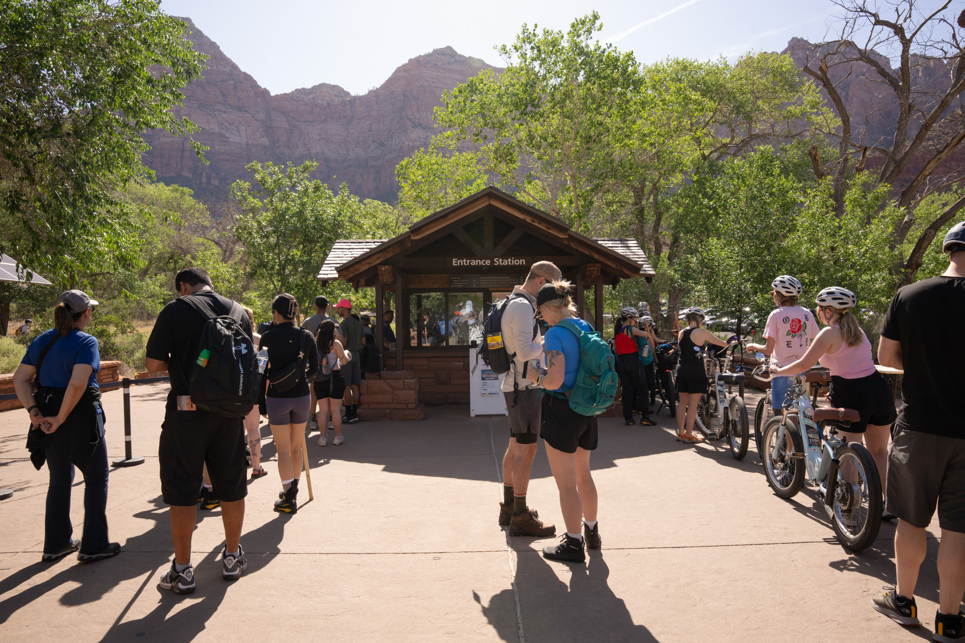 A small wooden building stands in a tree-lined plaza. On its sides, visitors form two lines. One side has visitors with bicycles