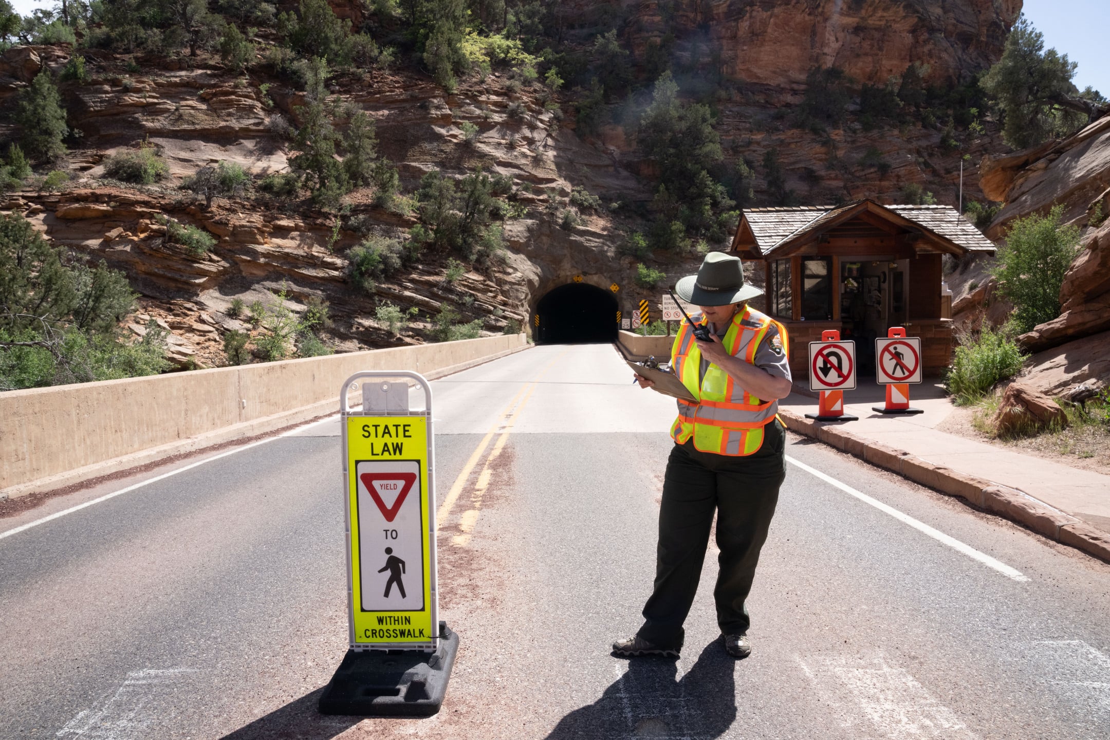 A park ranger in a high-visibility vest talks into a portable radio. In the background, a road leads into a sandstone tunnel.