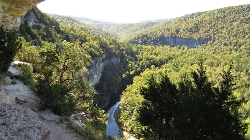 A narrow river can be seen flowing at the bottom of a canyon. Rolling, tree covered hillsides and sheer rock bluffs can be seen flanking both sides of the river corridor/NPS