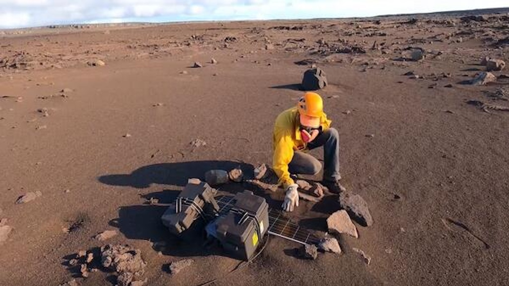 An HVO scientist clears tephra from a solar panel during the September 2021 Kīlauea summit eruption. A glove is used to brush away the Pele’s hair and other fine volcanic glass fragments to avoid small cuts and splinters. While working within the closed h An HVO scientist clears tephra from a solar panel during the September 2021 Kīlauea summit eruption. A glove is used to brush away the Pele’s hair and other fine volcanic glass fragments to avoid small cuts and splinters. While working within the closed h