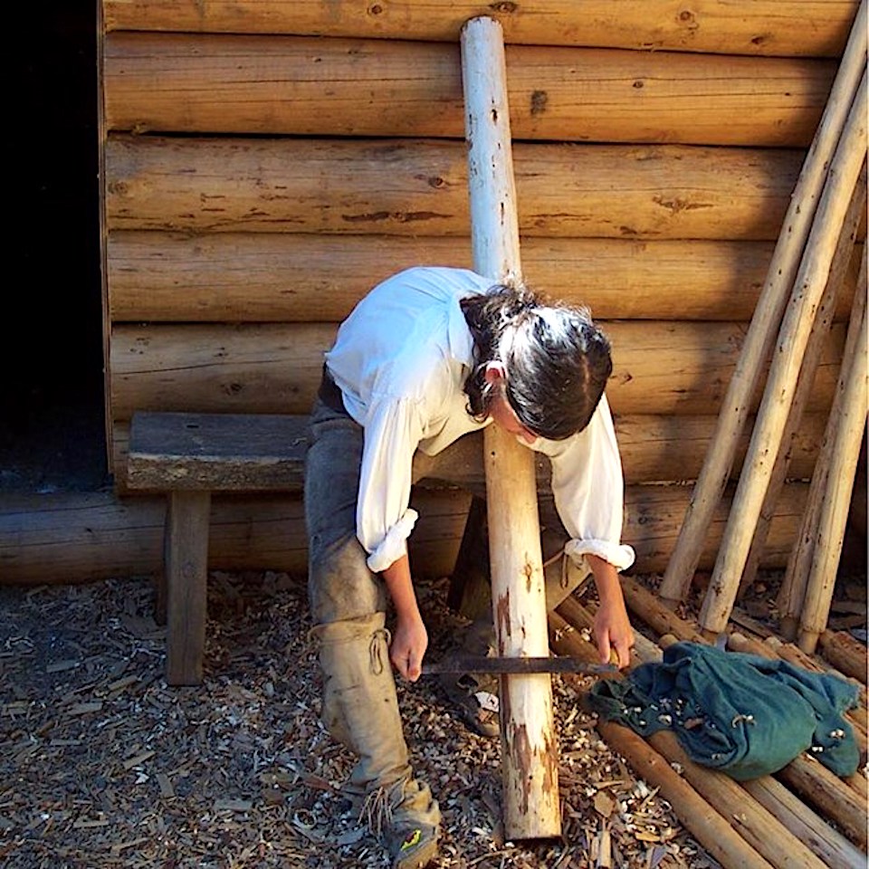 A park ranger, dressed in costume as Corps of Discovery member and carpenter Patrick Gass. scrapes wood off a tree pole. Gass was likely the carpenter in charge of building Camp Dubois, Fort Mandan, and Fort Clatsop/NPS
