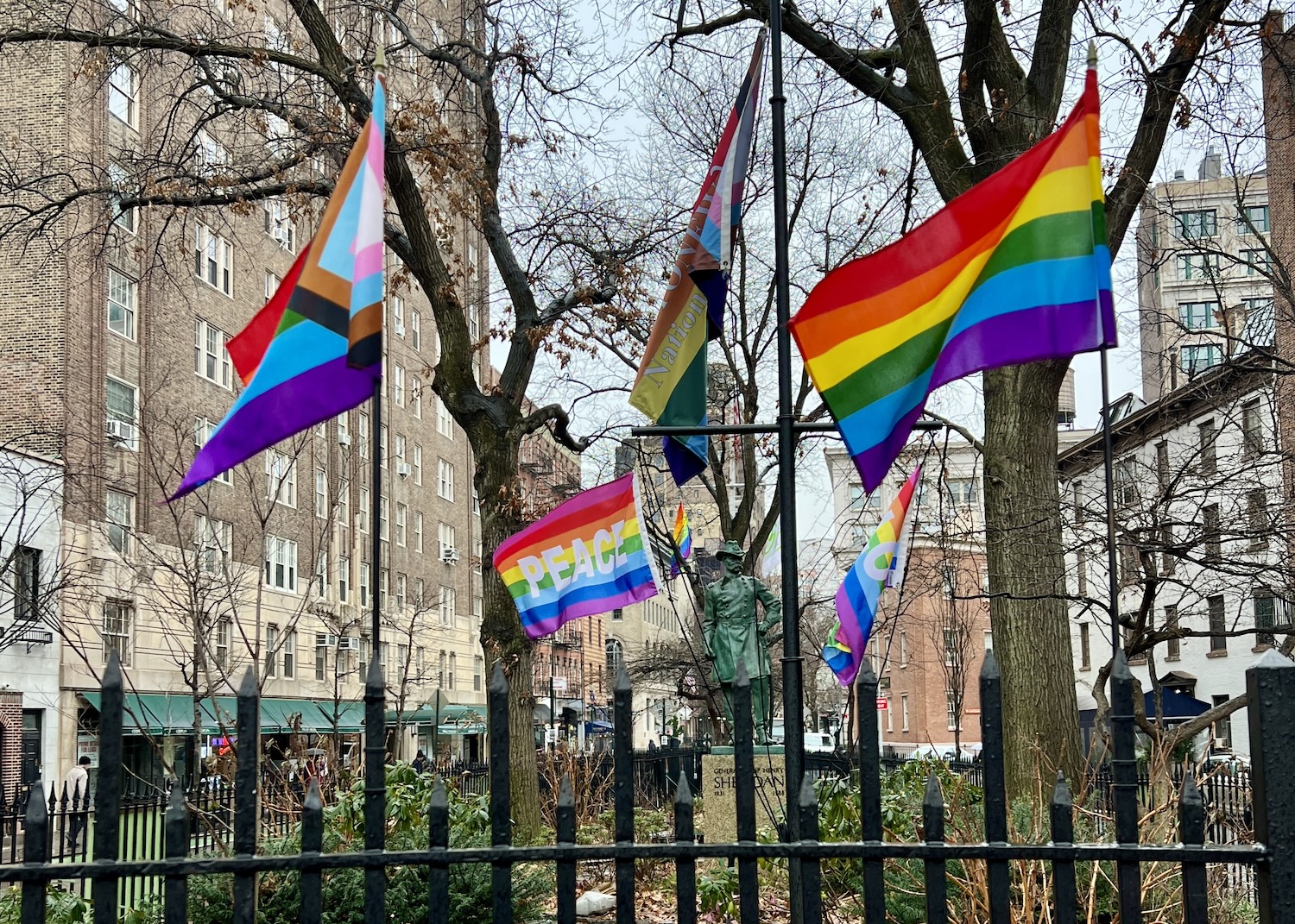 At Stonewall National Monument, various pride flags flutter in front of the statue of a Civil War general. At Stonewall National Monument, various pride flags flutter in front of the statue of a Civil War general.