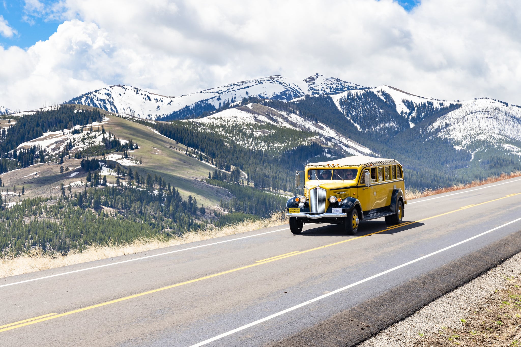 Dunraven Pass In Yellowstone National Park A yellow tour vehicle drives on a clear road surrounded by snow-capped mountains.