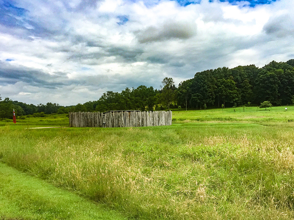 A small fort surrounded by tall green grass with a line of trees in the background beneath fluffy clouds in a blue sky, Fort Necessity National Battlefield.