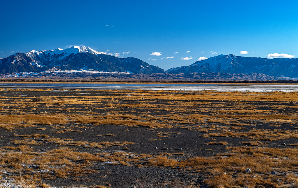 The view from San Luis Lakes State Wildlife Area toward a broad, flat valley below a distant dunefield and mountains beneath a blue sky, Great Sand Dunes National Park and Preserve