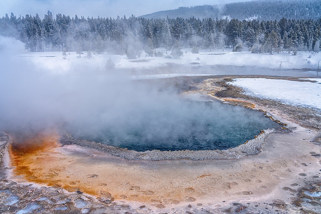 A telephoto view of Crested Pool at Upper Geyser Basin, with visible human and bison footprints along the edge of the hot spring in Yellowstone National Park