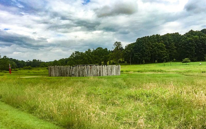 A small fort surrounded by tall green grass with a line of trees in the background beneath fluffy clouds in a blue sky, Fort Necessity National Battlefield.