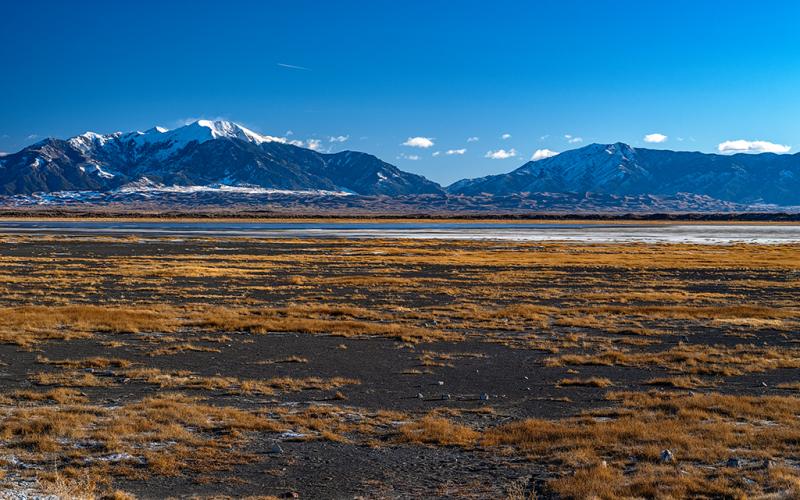 The view from San Luis Lakes State Wildlife Area toward a broad, flat valley below a distant dunefield and mountains beneath a blue sky, Great Sand Dunes National Park and Preserve