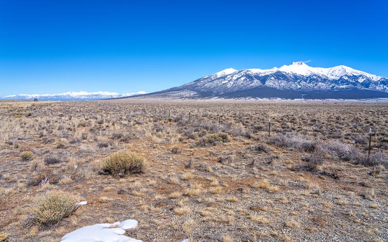 A vast panorama of road (CO Hwy 150) to the far left, a broad swath of scrub brush and San Luis Valley, and mountains (including Blanca Peak) to the right, highlighting a portion of the route of the Old Spanish National Historic Trail