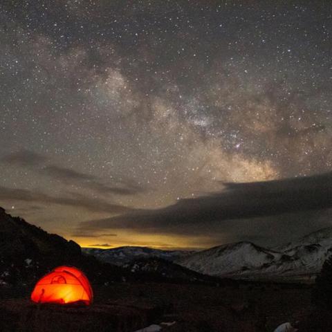 Orange light glows from a tent under a clear bright milky way blazing across the sky.
