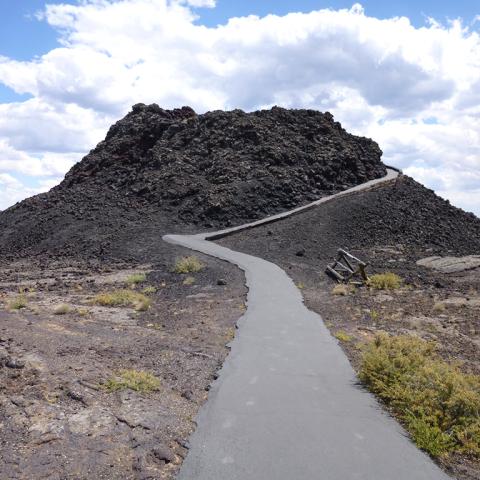 A small, black volcanic cone with a paved trail winding up and around the side, Craters of the Moon National Monument and Preserve