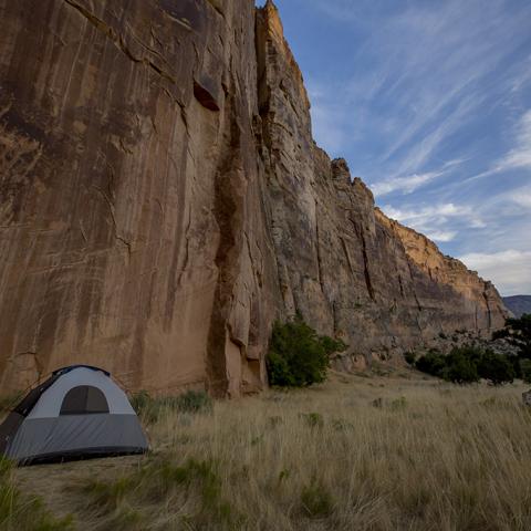 A tent set up against a high, red-rock cliff wall with a patch of blue sky and clouds overhead, Dinosaur National Monument