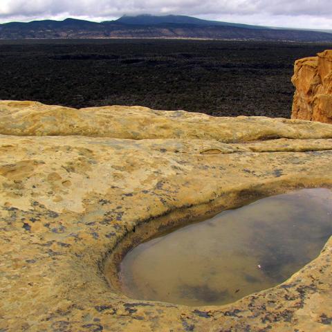 An oval-shaped depression filled with water rests in the surface of a tan cliff. Gray storm clouds fill the sky. Black lava flows below the cliffs appear flat. A distant mountain range lines the edge of the lava flows at El Malpais National Monument