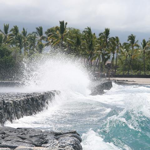 An overcast day and seaspray from a teal-colored ocean breaking along a stone wall at the Kaloko fish pond in Kaloko-Honokohau National Historical Park