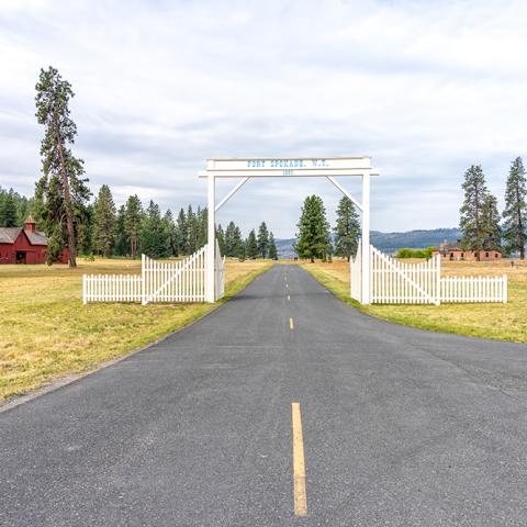 A road leading beyond a white entrance gate with blue lettering, and a field of grass on either side of the road. The red-painted wooden Quartermaster's Stable sits on the left side of the road beyond the gate at Fort Spokane in Lake Roosevelt National Recreation Area.