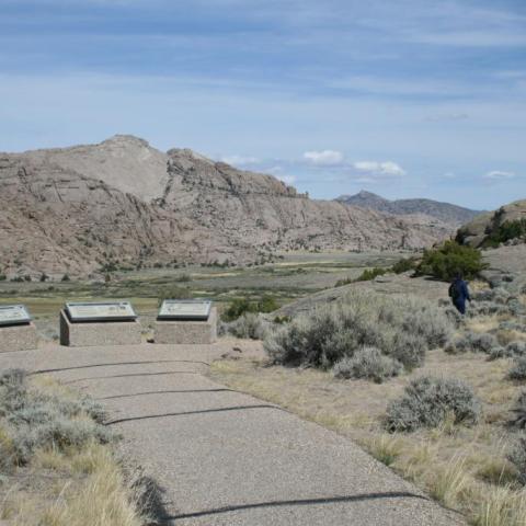 Two visitors walk on a trail facing a large brown rock buttress, a paved path leads to three exhibits.