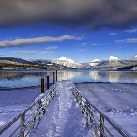 A snow-covered leading line of the dock at Lake McDonald leading the viewer's eye across the water and toward the distant, snow-covered mountains at Glacier National Park