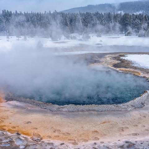 A telephoto view of Crested Pool at Upper Geyser Basin, with visible human and bison footprints along the edge of the hot spring in Yellowstone National Park