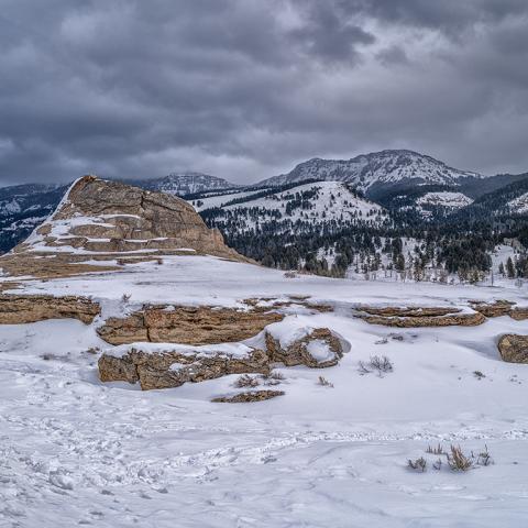 Soda Butte travertine cone on a snowy winter day, with the Absaroka Mountains in the background, all beneath a sky filled with dark, stormy clouds in Yellowstone National Park.