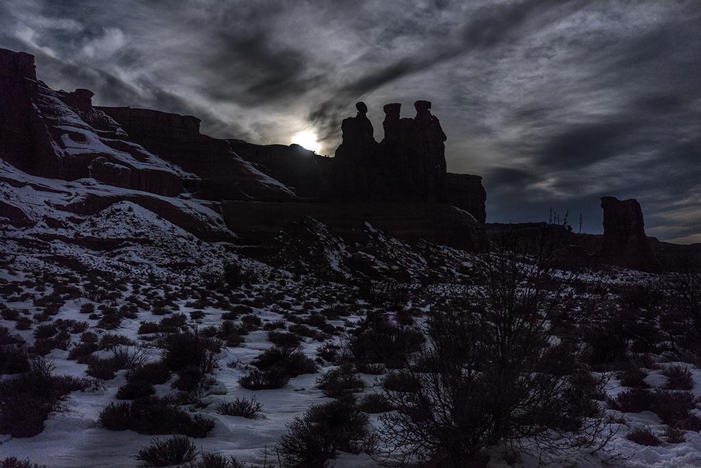 A full moon on a cloudy, snowy night, silhouetting the Three Gossips stone formation in Arches National Park, in Utah