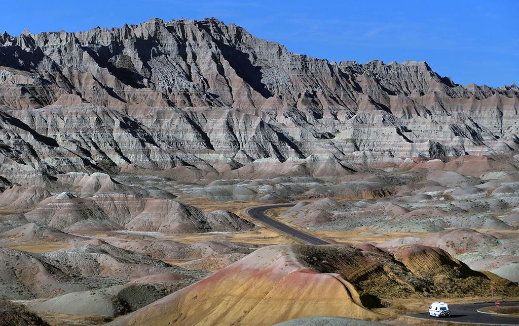 A camper on a paved park road dwarfed by immense, surreal, layered geological formations in South Dakota's Badlands National Park