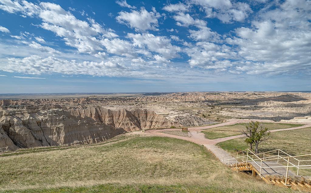 A blue sky with puffy white clouds over a basin of layered badlands formations and a grassy area with a fenced overlook looking out to the basin, Badlands National Park, South Dakota