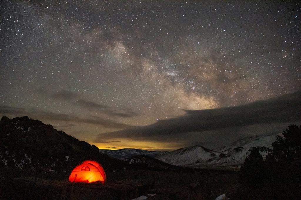 Orange light glows from a tent under a clear bright milky way blazing across the sky.