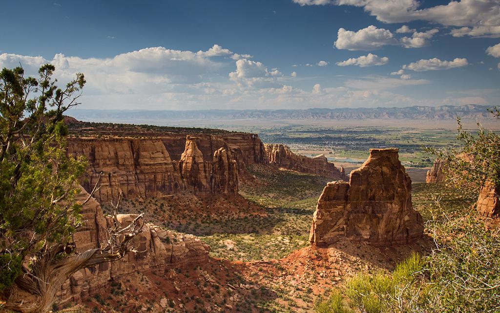 Red-rock formations with the town of Grand Junction far in the distance, and mountains even further in the background, all under a hazy blue sky with clouds, Colorado National Monument