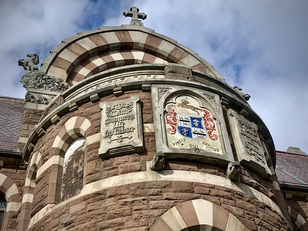 Brown and white stone building with a round portion with long curved windows with a blue and red crest in the middle. A cross sits atop the arch with a colt flanking each side, Coltsville National Historical Park