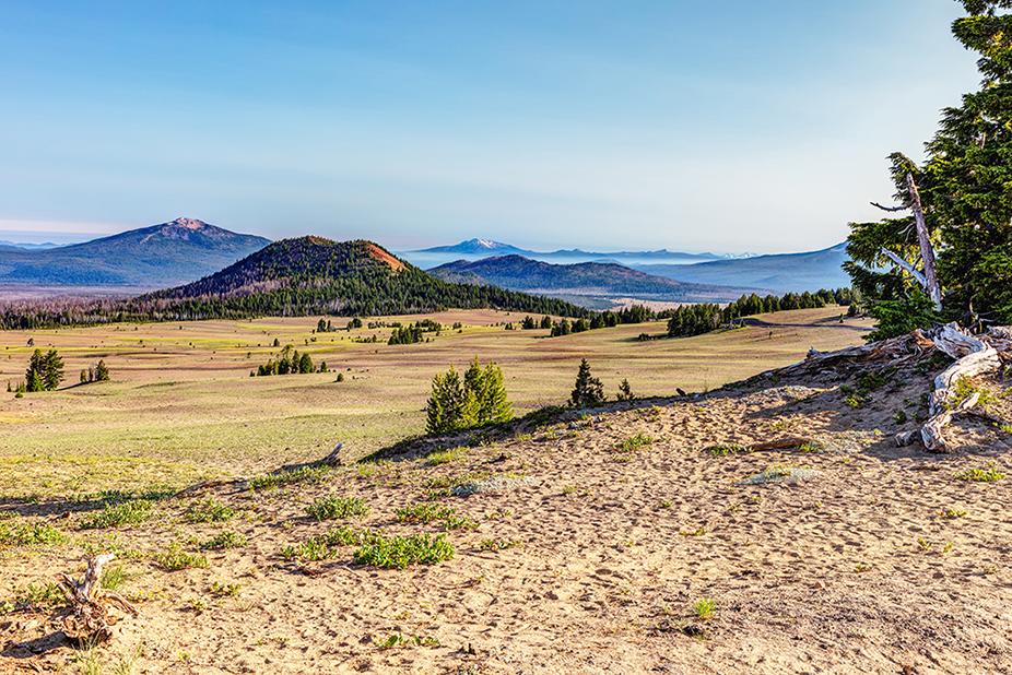A view of distant mountains and a tree-covered cinder cone on a wide expanse of volcanic field in Crater Lake National Park, Oregon