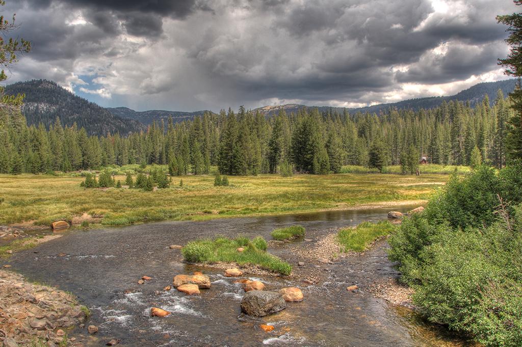 A shallow river bordered by bright green meadow and trees beneath a moody, cloudy sky, Devils Postpile National Monument