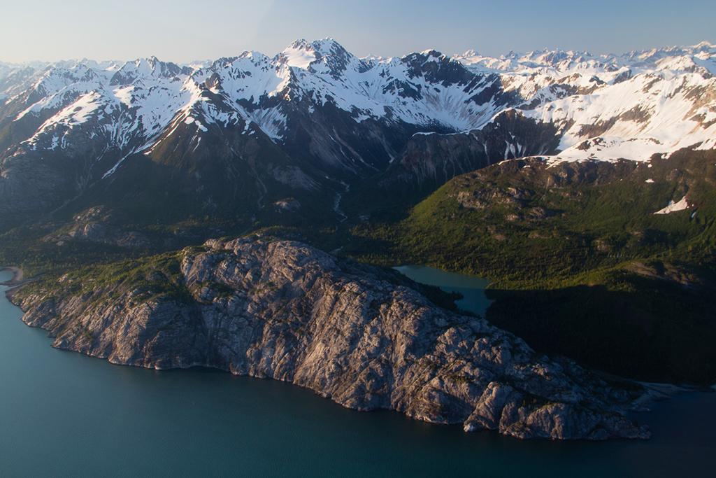 An aerial view of large, bare, rocky mountain against backdrop of white capped mountains of Gloomy Knob, Glacier Bay National Park and Preserve