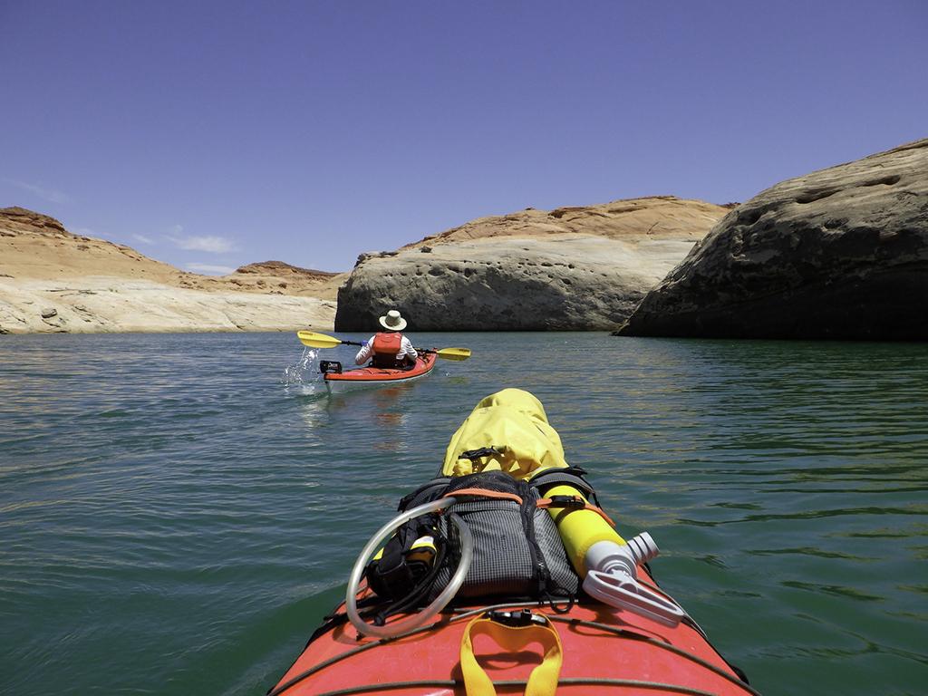 A view from one kayak looking toward another kayaker on green water looking toward red rocks in Hansen Canyon on Lake Powell at Glen Canyon National Recreation Area