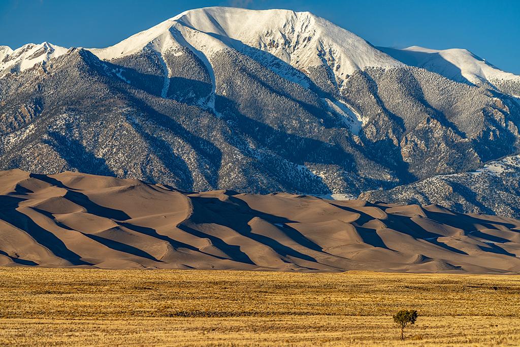 A telephoto view of a snowcapped mountain towering over a dunefield sitting on a flat expanse of land, Great Sand Dunes National Park and Preserve