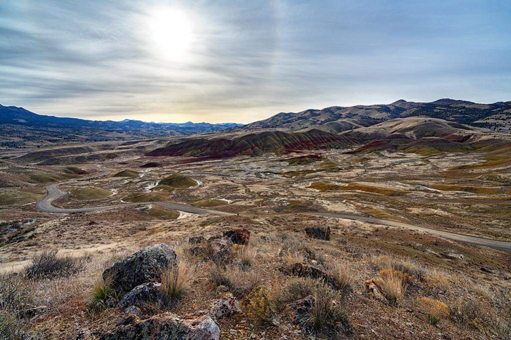 A sun halo over an expansive view of hills layered in colors of beige, maroon, and yellow-green seen from the top of the Carroll Rim Trail in the Painted Hills Unit of John Day Fossil Beds National Monument.