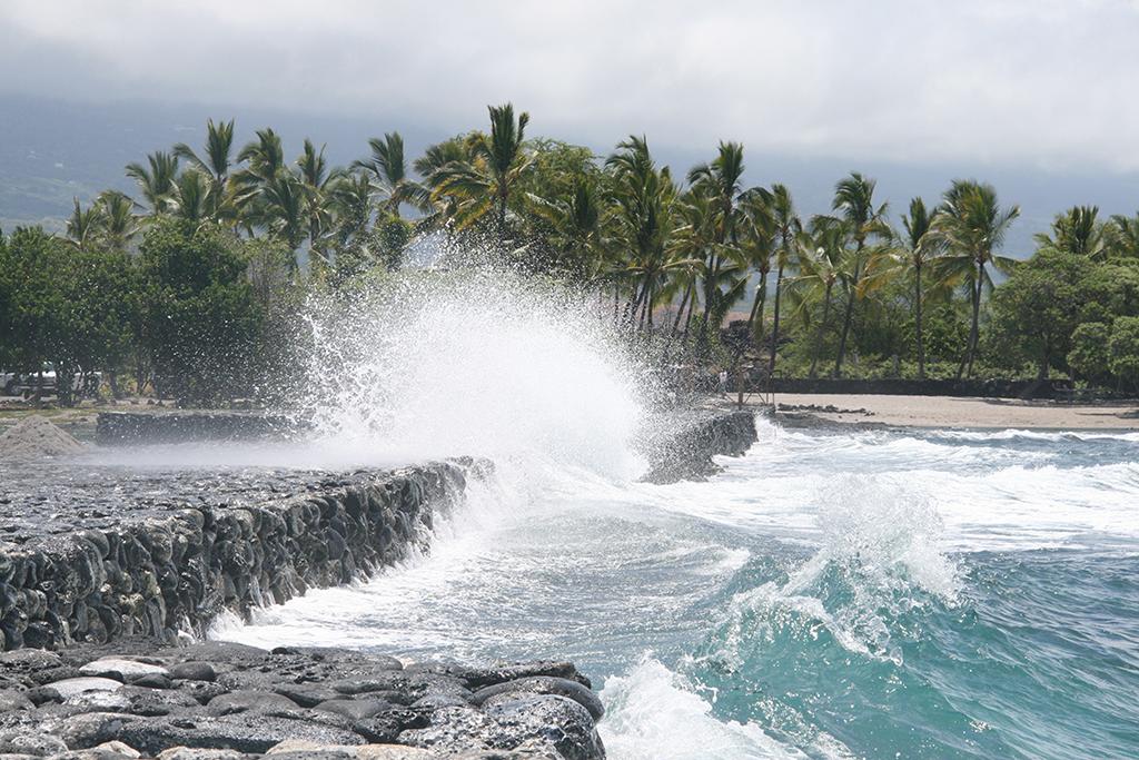 An overcast day and seaspray from a teal-colored ocean breaking along a stone wall at the Kaloko fish pond in Kaloko-Honokohau National Historical Park