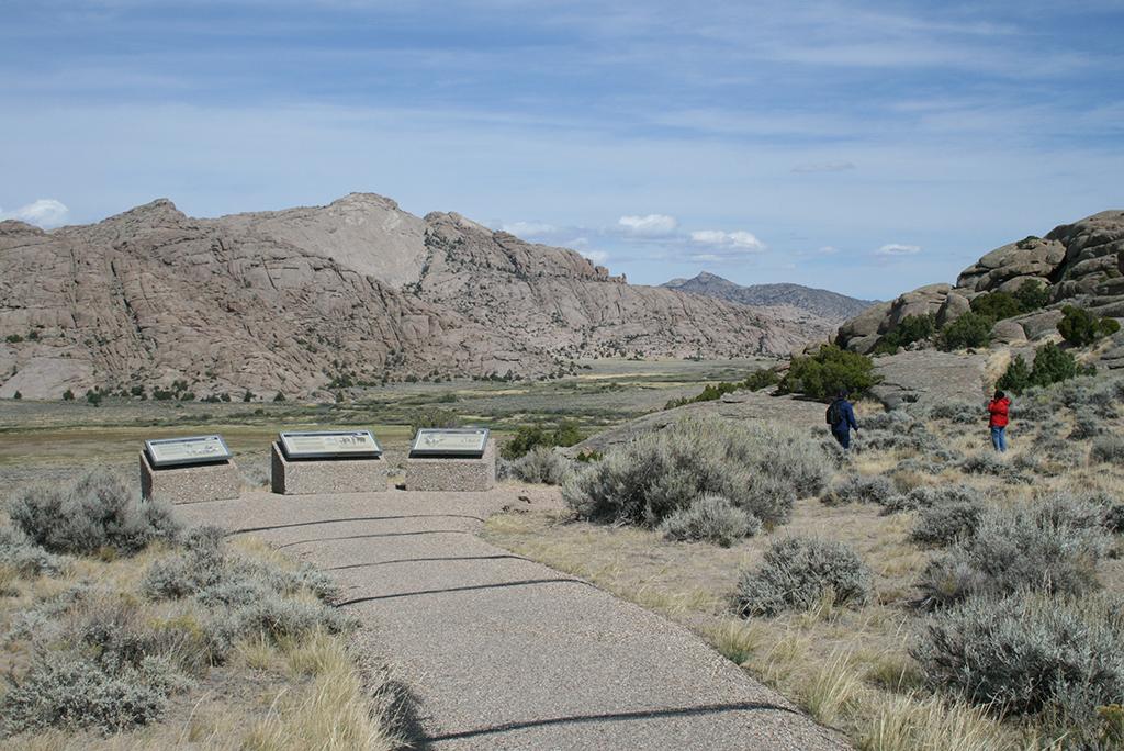 Two visitors walk on a trail facing a large brown rock buttress, a paved path leads to three exhibits at Split Rock Viewpoint, Mormon Pioneer National Historic Trail