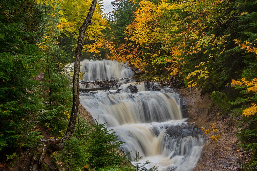 The silky white and brown water of Sable Falls framed by green and autumn-gold trees at Pictured Rocks National Lakeshore in Michigan