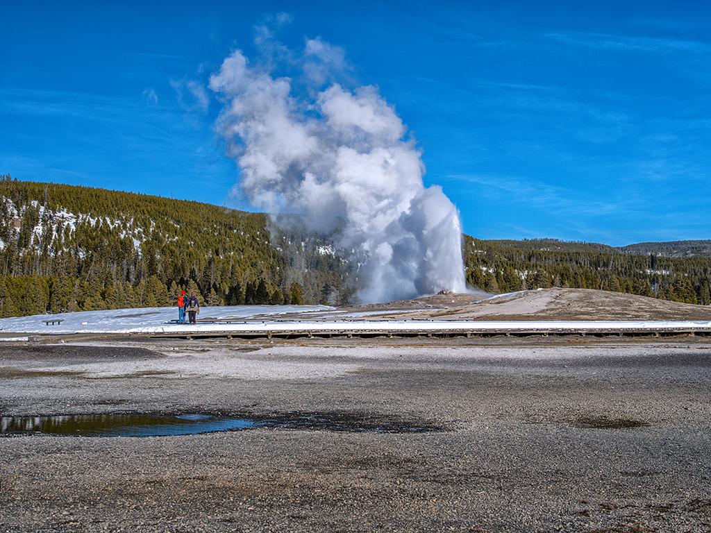 Two people standing together, watching Old Faithful Geyser erupt on a blue-sky winter day in Yellowstone National Park