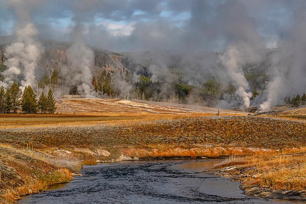 an image of the Firehole River flowing between a golden landscape with steaming geysers and hot springs at Upper Geyser Basin in Yellowstone National Park