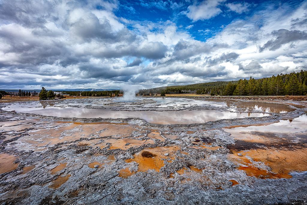 A wide-angle view of a quiet Great Fountain Geyser, with reflections in the calm water and puffy clouds in a blue sky at Yellowstone National Park