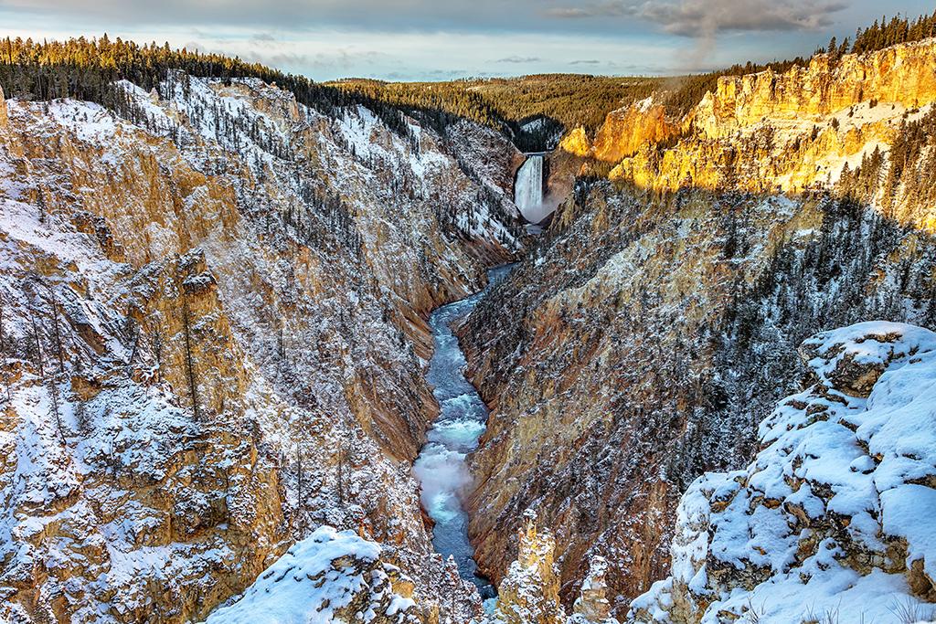 A view of the Lower Falls of the Yellowstone River with sunlight turning the upper right portion of the canyon a golden yellow while the rest of the area is shaded and blanketed with light snow and frost, Yellowstone National Park