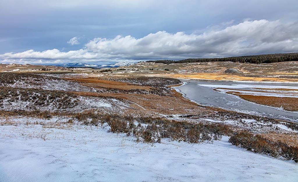 A snow-dusted wide-angle view of Hayden Valley on a frosty autumn day, with puffy clouds in the sky and rugged mountains in the distance in Yellowstone National Park