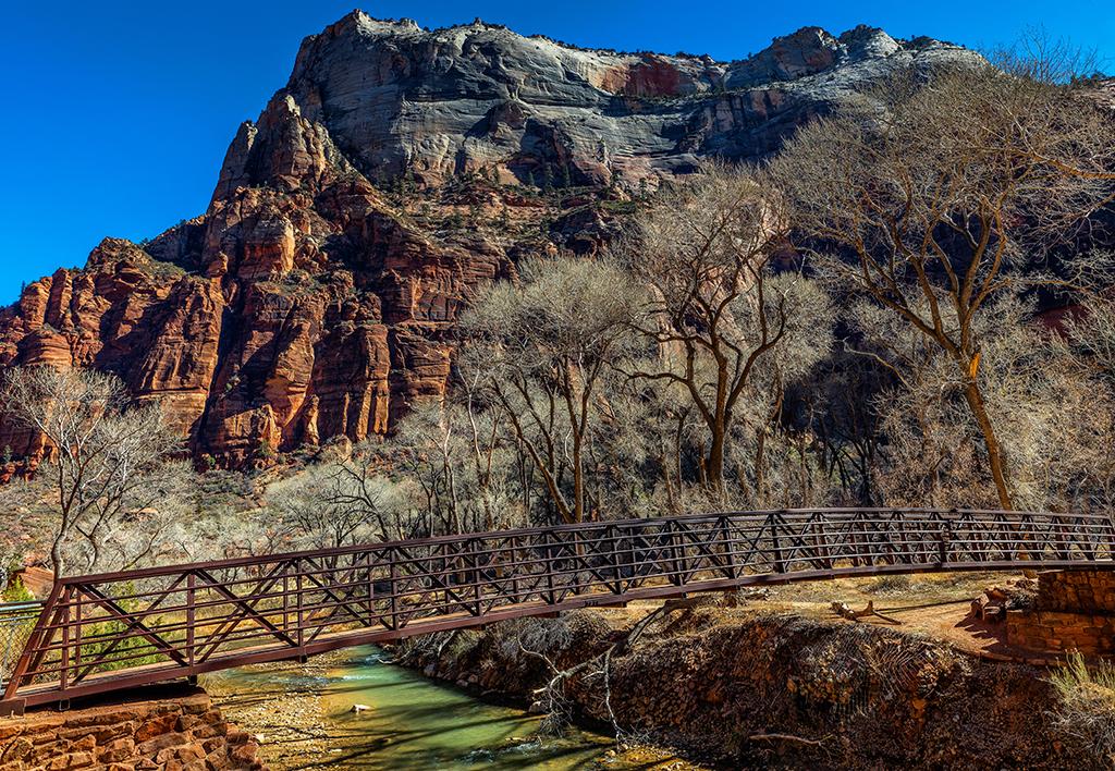 A view of the bridge spanning the Virgin River, connecting the park road with the Angels Landing Trail, with tall canyon walls in the background and bare tree branches in Zion National Park