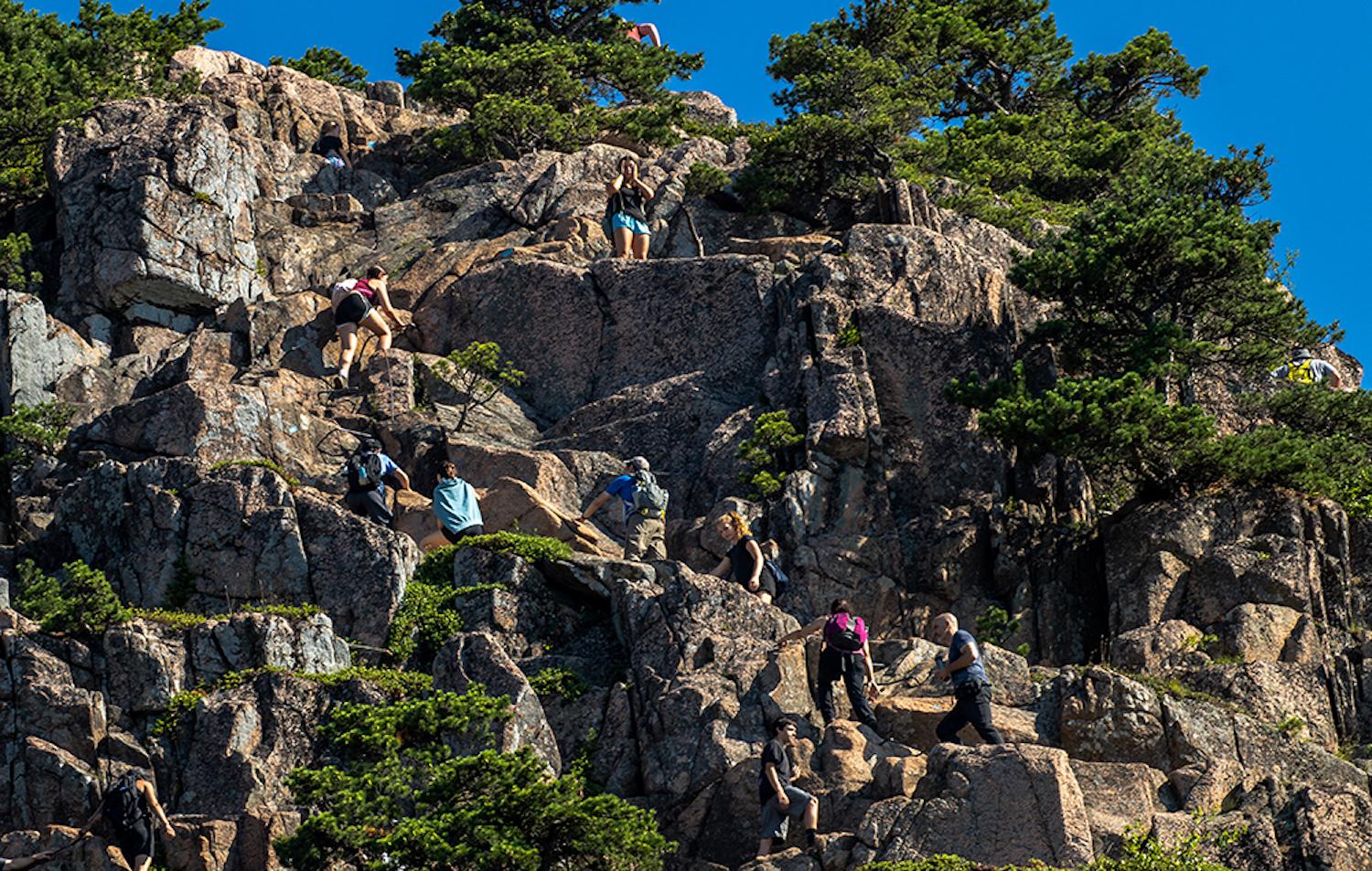 A line of hikers climb up the Beehive Trail on Tuesday, June 29, 2021 in Acadia National Park. As the park’s volume of visitors increase and the summer season gets hotter, more and more visitors are trekking the more popular trails as early as possible – 